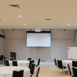 A conference room set up with round tables covered in white cloths and black chairs facing a large projector screen on the stage area.