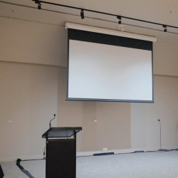 Mobile presentation lectern with microphone in front of a large pull-down projection screen in a conference room edge of a stage area
