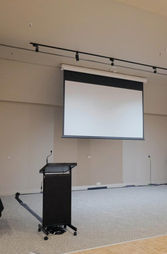 Mobile presentation lectern with microphone in front of a large pull-down projection screen in a conference room edge of a stage area