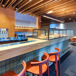 Pizza shop interior with a glass display case of pizzas along a wooden counter and orange bar stools in the foreground.