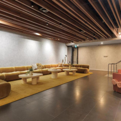 Modern lounge with a long mustard sofa, round coffee tables, warm lighting, and a wood-slatted ceiling; pink seating near the stairs.