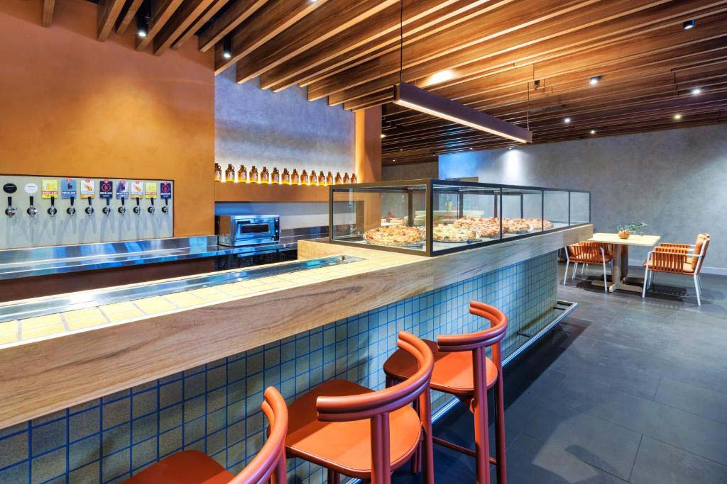 Pizza shop interior with a glass display case of pizzas, beer taps along a counter, and orange bar stools at a tiled front edge.