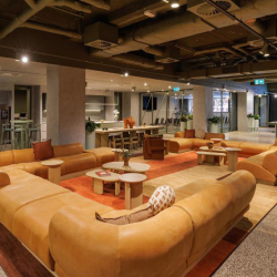 Open-plan lounge with a large orange sectional sofa forming a circle around small coffee tables on a rug; kitchen area visible in the background under an exposed ceiling.