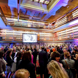 Crowded social event in a grand library-like room with multiple levels of bookshelves and a projector screen at the center.