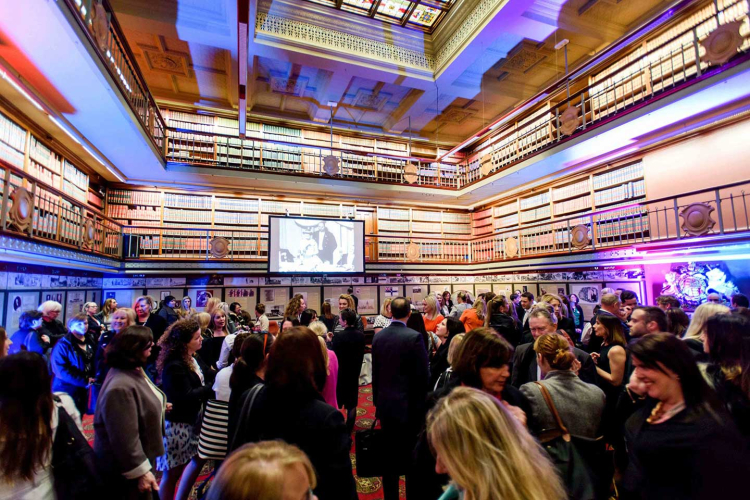 Crowded social event in a grand library-like room with multiple levels of bookshelves and a projector screen at the center.