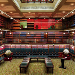Grand early 20th-century library reading room with two levels of bookshelves, globe displays, and ornate railings surrounding a central carpeted floor.