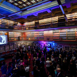 Crowded reception in a grand library with balconies, towering bookshelves, and a large screen showing a black-and-white image.