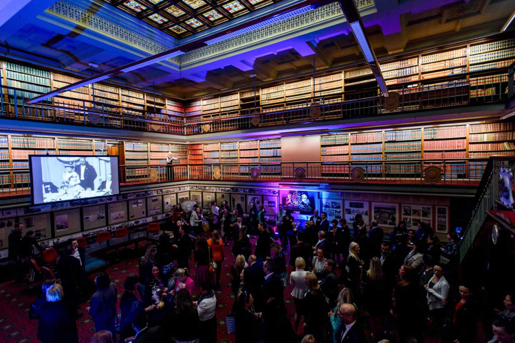 Crowded reception in a grand library with balconies, towering bookshelves, and a large screen showing a black-and-white image.