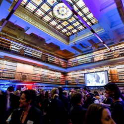 Guests mingle at a formal event in a grand library with a stained glass skylight and tall bookshelves, a projector screen in the background.