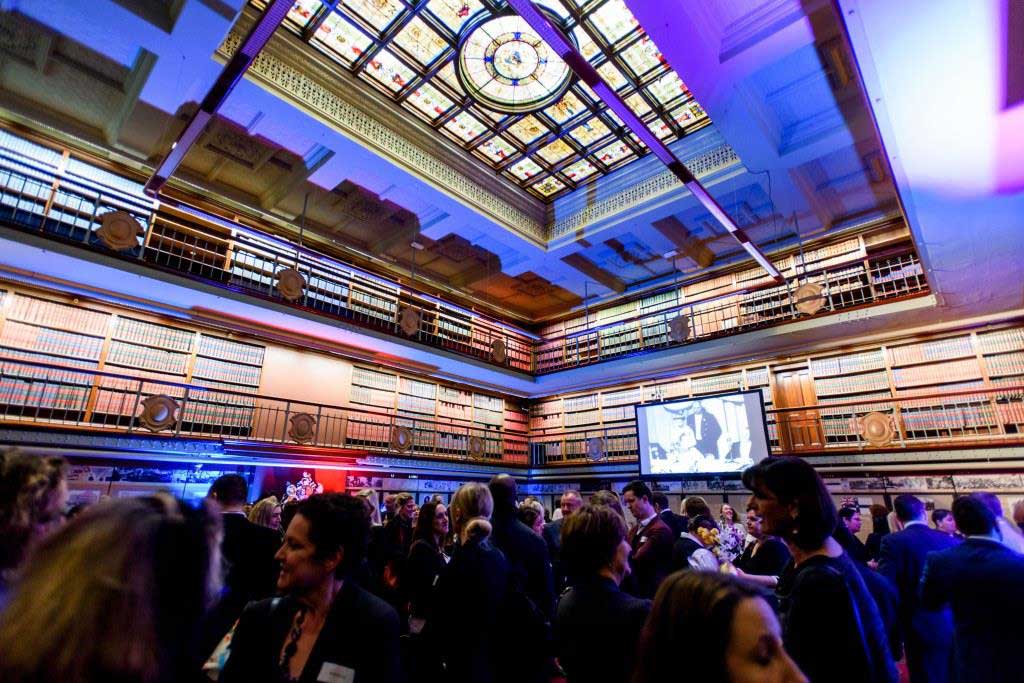Guests mingle at a formal event in a grand library with a stained glass skylight and tall bookshelves, a projector screen in the background.