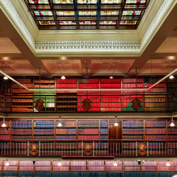 Interior of a grand, multi-level library with balconies, ornate railings and a colorful stained-glass skylight above.