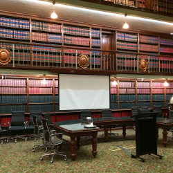 Historic library conference room with tall wooden bookshelves, a projection screen, a globe, and rows of black rolling chairs around dark wooden tables.