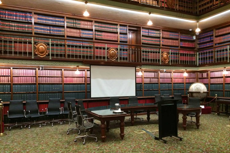 Historic library conference room with tall wooden bookshelves, a projection screen, a globe, and rows of black rolling chairs around dark wooden tables.