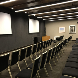 Conference room set with multiple rows of black chairs facing a front stage and white projection screens on dark walls, framed photos on the side wall.