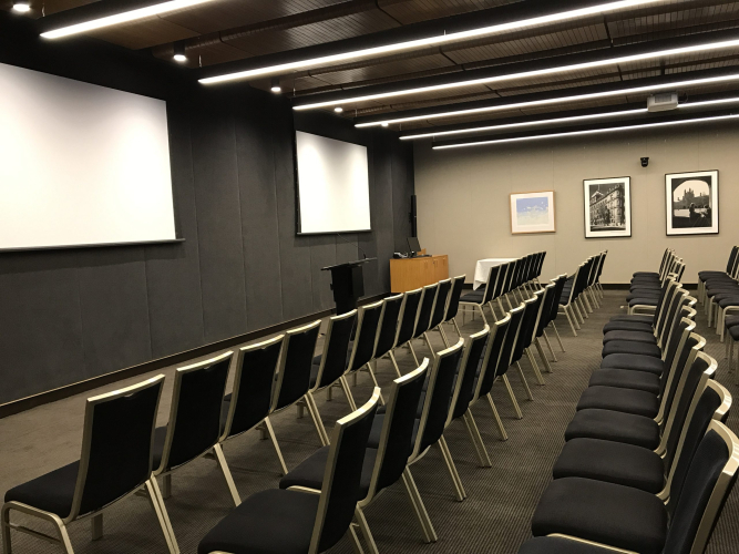 Conference room set with multiple rows of black chairs facing a front stage and white projection screens on dark walls, framed photos on the side wall.