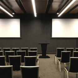Empty conference or lecture room set up for a presentation: two large white projection screens on a dark wall, a black podium at center, and rows of black chairs facing them.