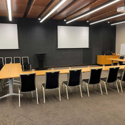 A conference room set up in a U-shaped desk arrangement with black chairs and water glasses, two large projection screens, and a podium at the front.