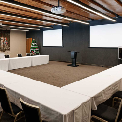 U-shaped conference room setup with white-tablecloth tables, a black podium, two blank projector screens, and a Christmas tree in the corner.