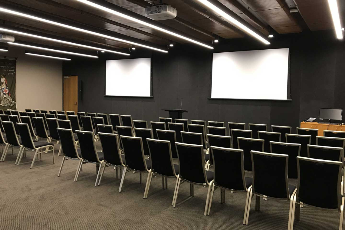 Empty conference room with two projection screens, a podium, and rows of black chairs facing the front.