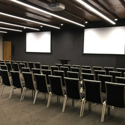 Empty conference room with two projection screens, a podium, and rows of black chairs facing the front.