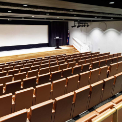 empty auditorium with a wooden stage, a large projection screen, a black curtain backdrop, a podium, and rows of brown upholstered seats; an Australian flag beside the stage