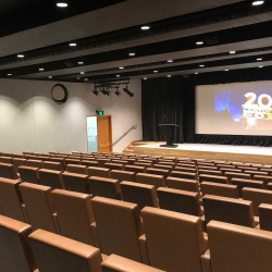 Empty theater with brown upholstered seats facing a stage and a large screen showing the 20th Century Fox logo.