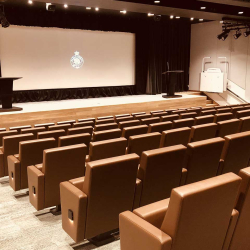 Empty lecture hall with rows of brown seats facing a stage and a large screen.