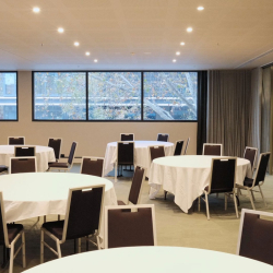 Conference room with round tables dressed in white cloths and brown chairs, large windows and soft lighting.