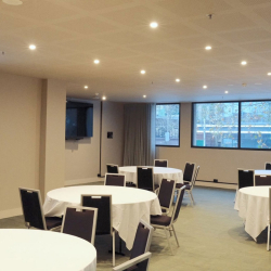Bright conference room set up for a reception with round tables, white tablecloths, and dark chairs; large windows overlook trees outside.