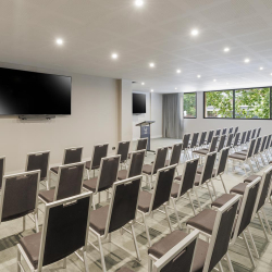 Conference-style room with rows of gray-cushioned chairs facing a lectern and two large wall-mounted TVs, and large windows overlooking greenery.