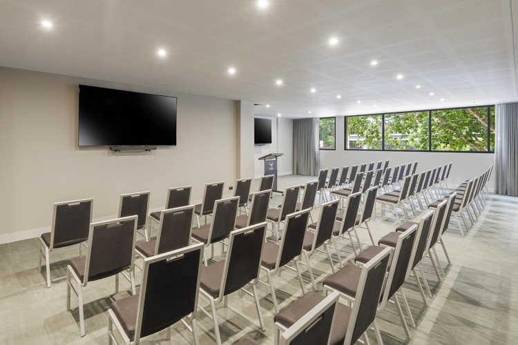 Conference-style room with rows of gray-cushioned chairs facing a lectern and two large wall-mounted TVs, and large windows overlooking greenery.