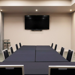 Conference room with a long, dark table and black chairs arranged for a meeting, TV mounted on a beige wall at the far end, soft ceiling lights.