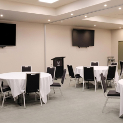 Empty conference room with round tables covered in white cloths, black chairs, a podium at the front, and two wall-mounted TVs.