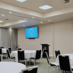 Conference room set up for a seminar with round tables draped in white cloths and black chairs around them, a lectern at the front, and a blue welcome screen on the wall.